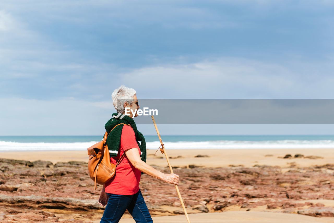 Side view of elderly female backpacker with trekking pole strolling on boulders against stormy ocean under cloudy sky