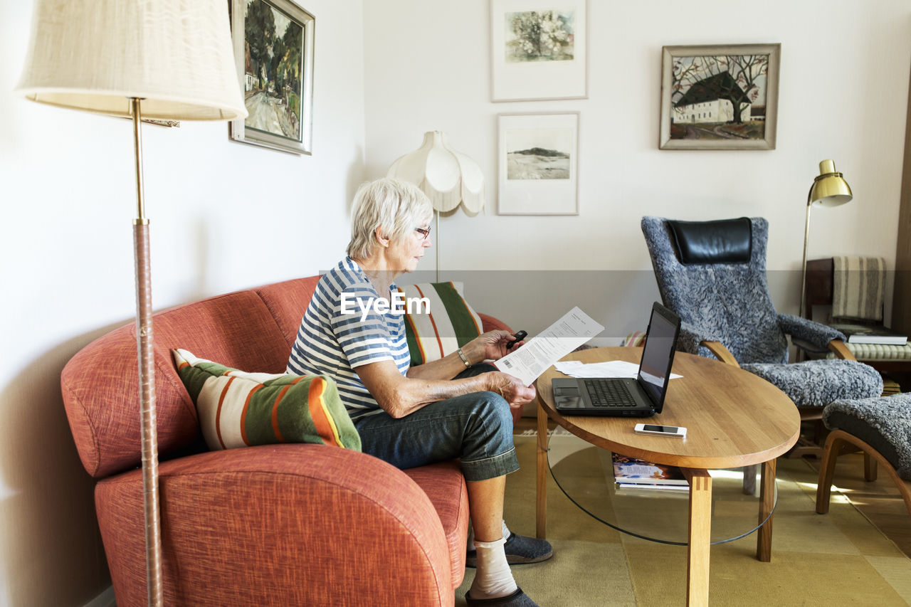 Senior woman analyzing document while using laptop at home