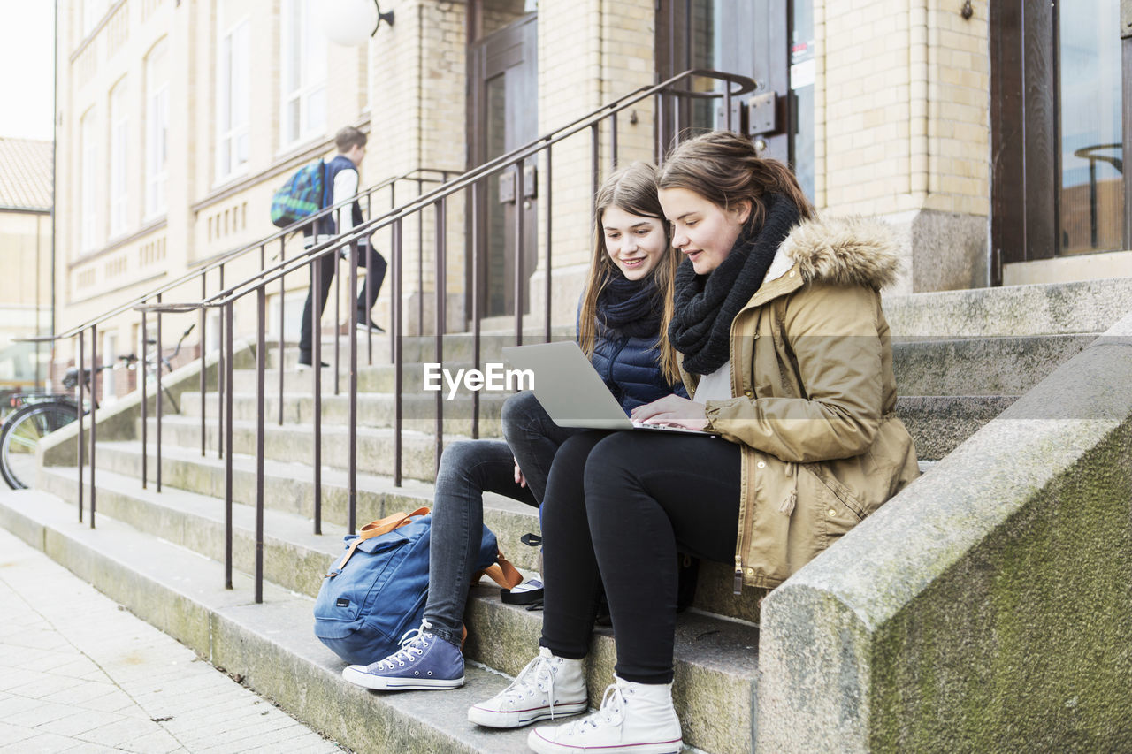 Young female friends using laptop on steps outside school building
