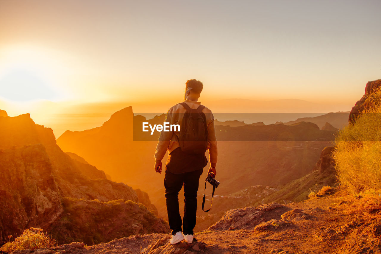 Rear view of man holding camera while standing on mountain against sky during sunset