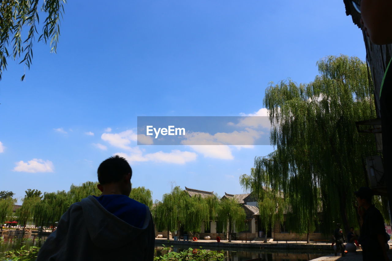 REAR VIEW OF MAN STANDING IN PARK AGAINST SKY