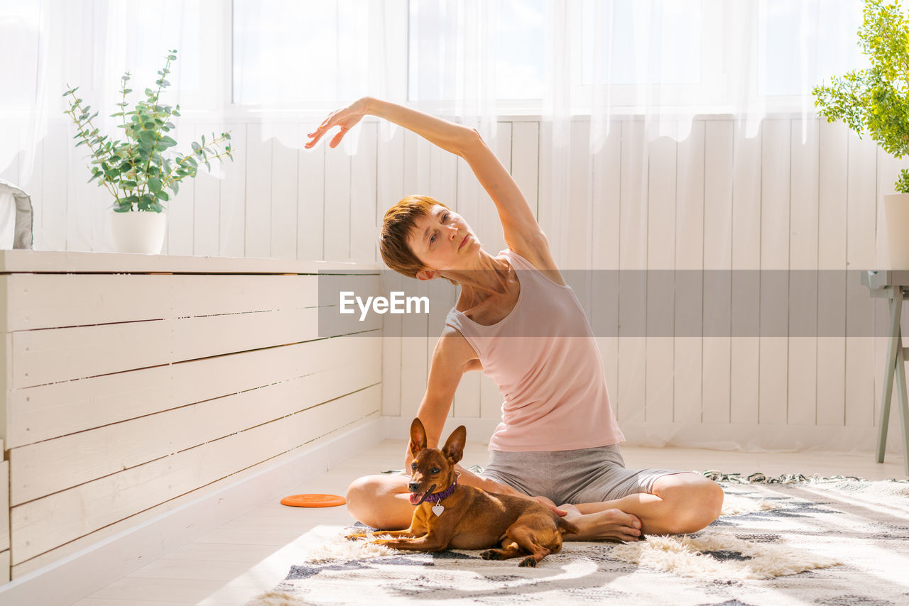 Beautiful adult woman practicing yoga pose with pygmy pinscher dog enjoying