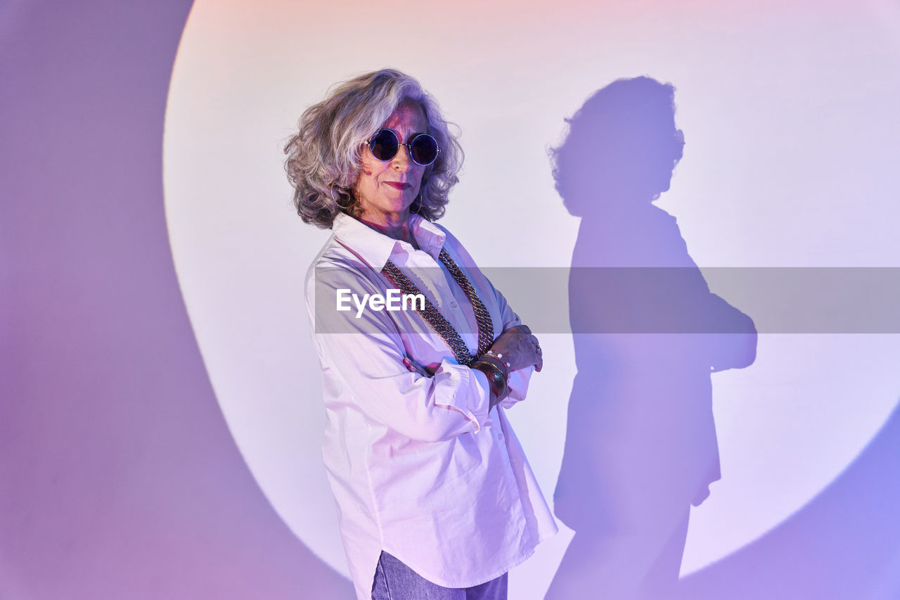Elegant elderly female in white shirt and sunglasses crossing arms while standing in bright spotlight circle and looking at camera