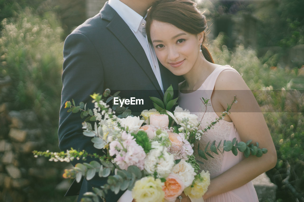 Portrait of bride holding bouquet while embracing groom against plants