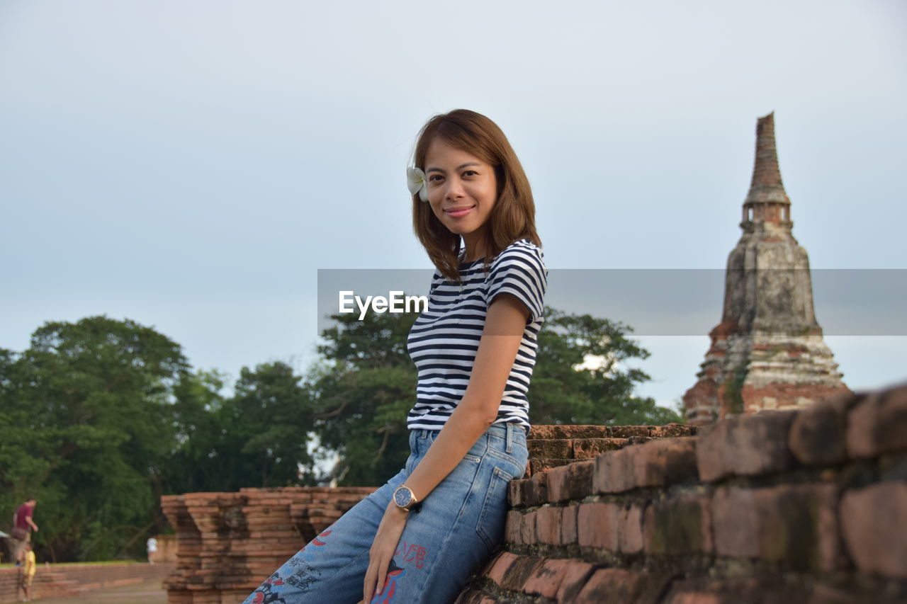 Portrait of woman leaning on brick wall against old temple in city