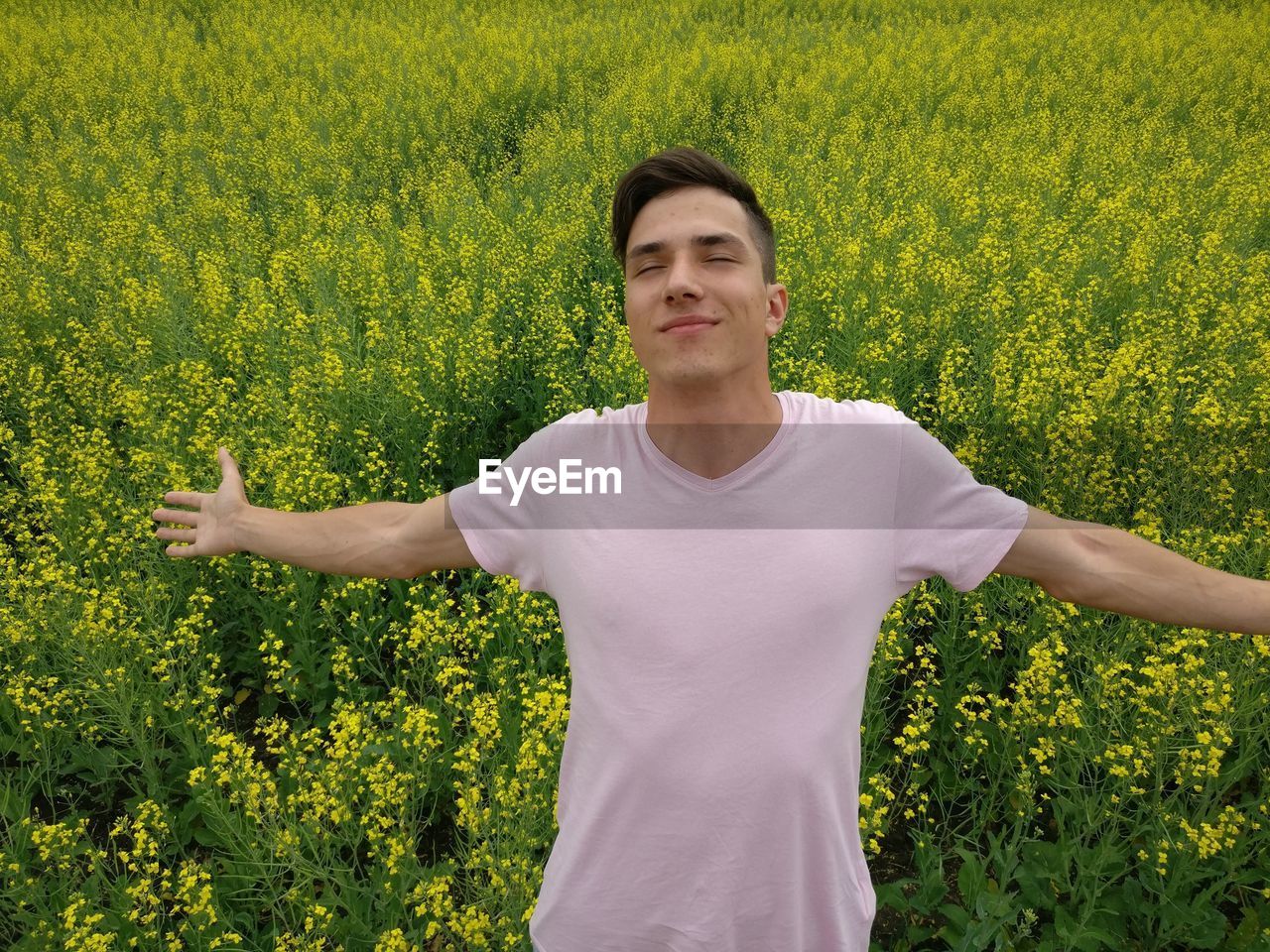 Smiling young man with eyes closed and arms outstretched standing against flowering plants at farm