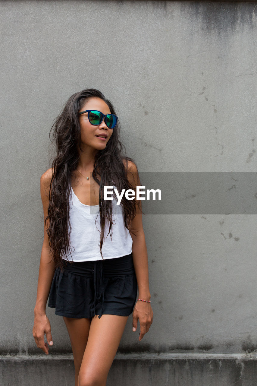 Young woman in sunglasses standing against wall