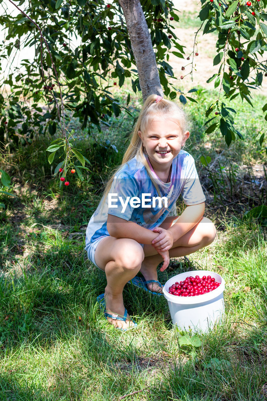 A happy girl smiles and squats next to a bucket of ripe cherry berries on a sunny summer day 
