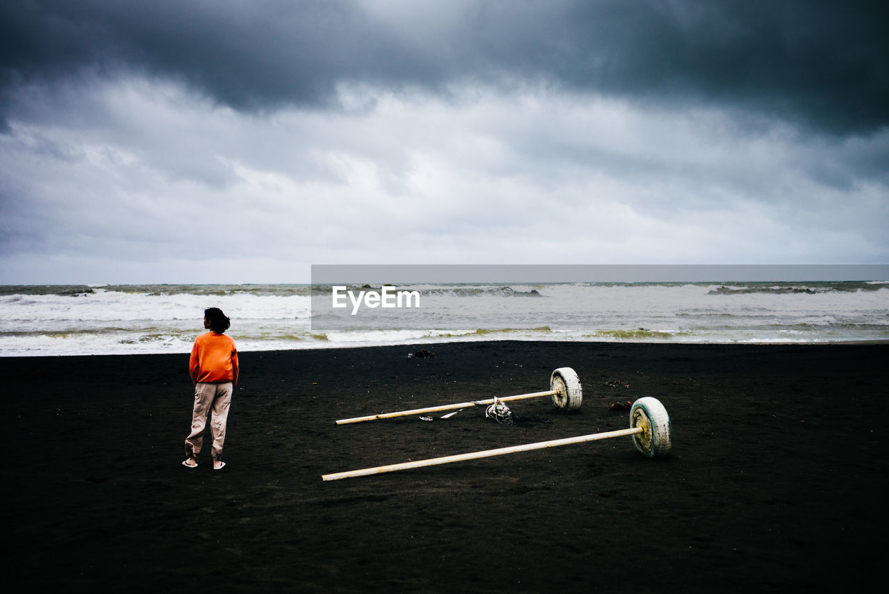 REAR VIEW OF MAN STANDING AT BEACH AGAINST SKY