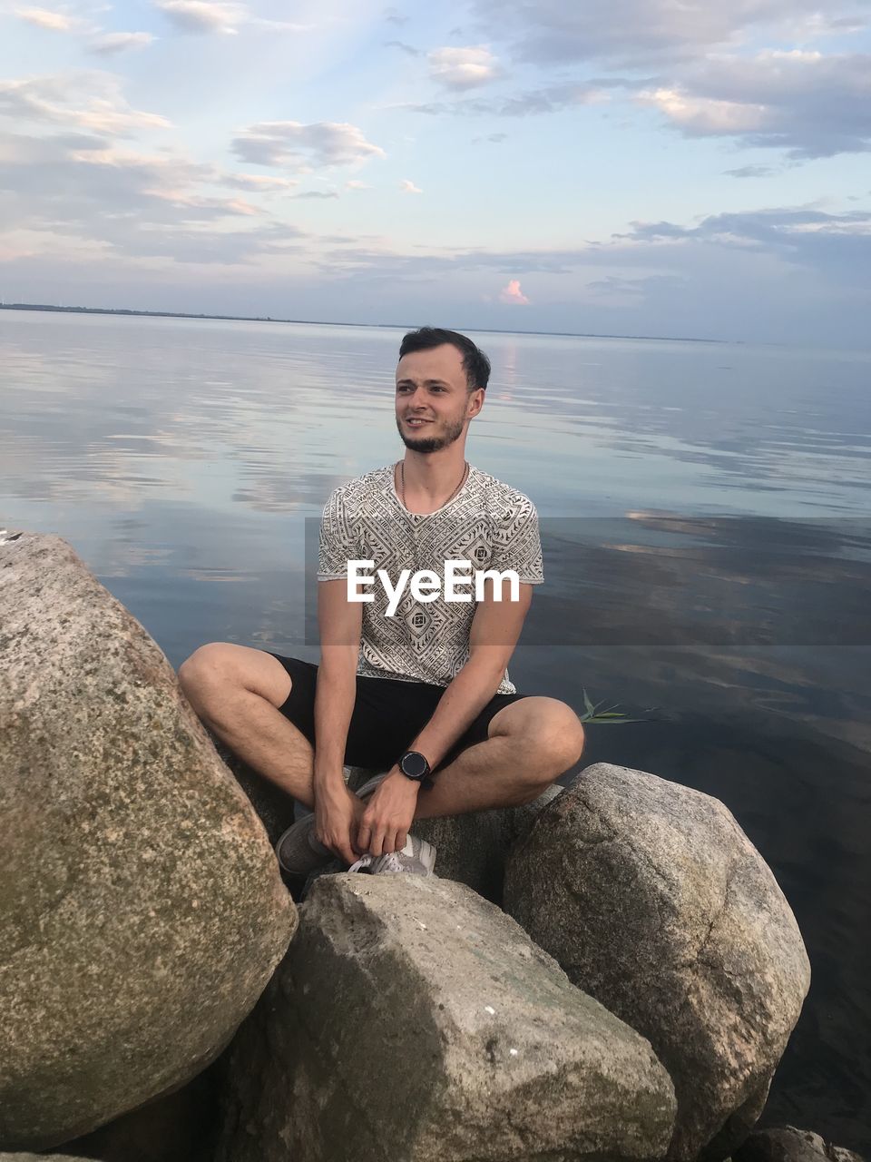 FULL LENGTH OF MAN SITTING ON ROCK AT SEA SHORE AGAINST SKY