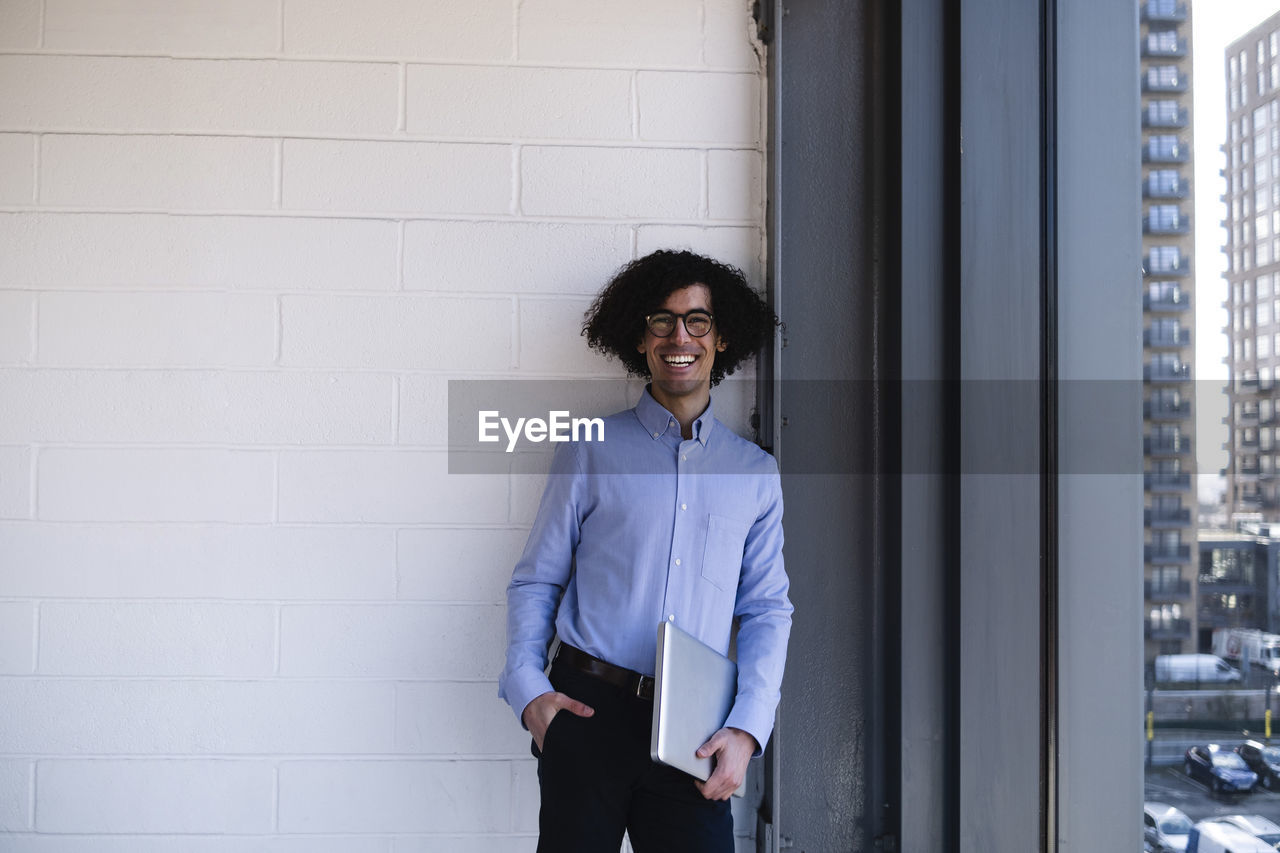 Happy businessman holding laptop standing with hand in pocket in office