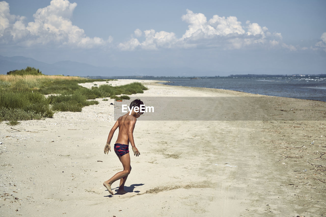 Side view of shirtless boy walking at beach against sky during sunny day
