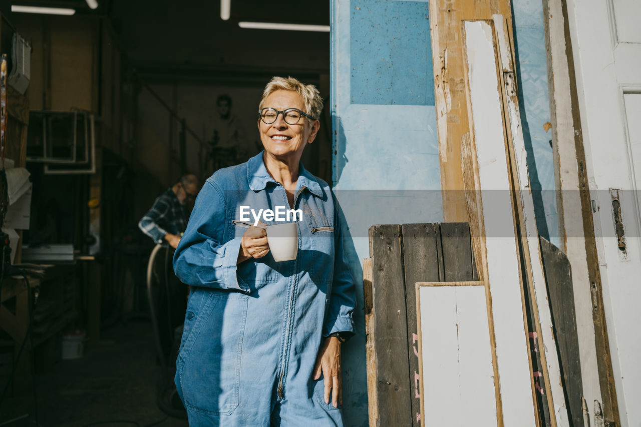 Happy female entrepreneur holding cup while standing near workshop doorway