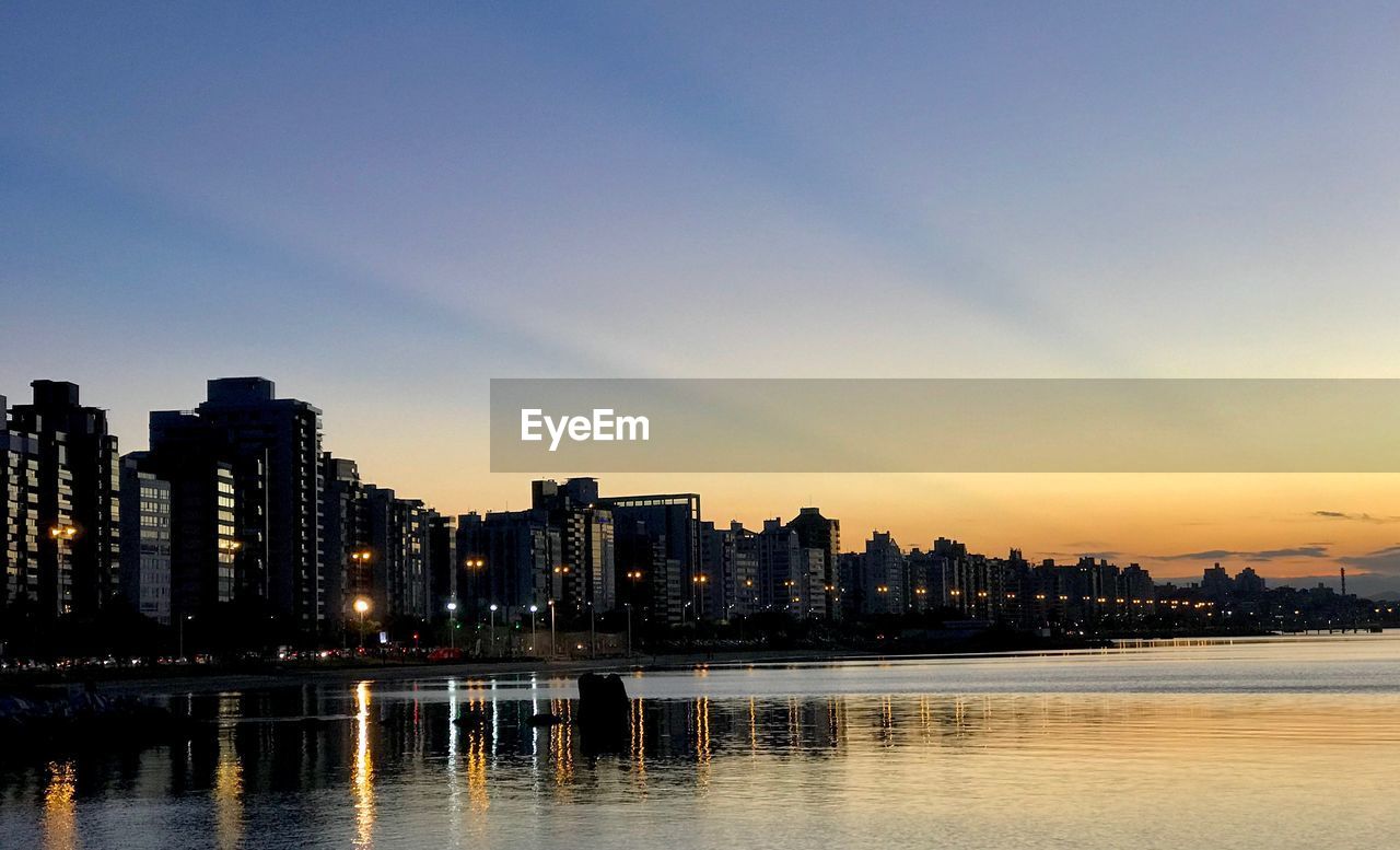 River by illuminated buildings against sky during sunset