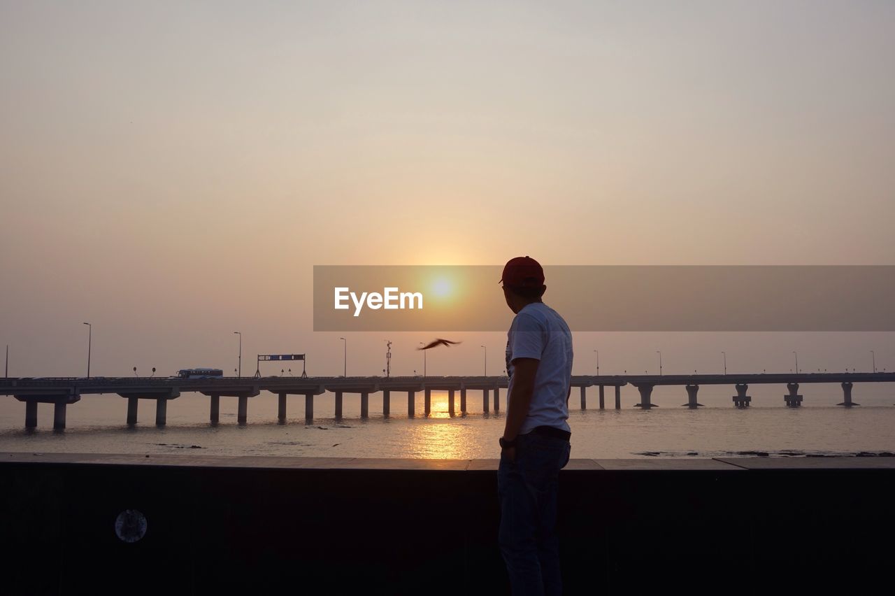 Side view of man standing by railing against sea during sunset