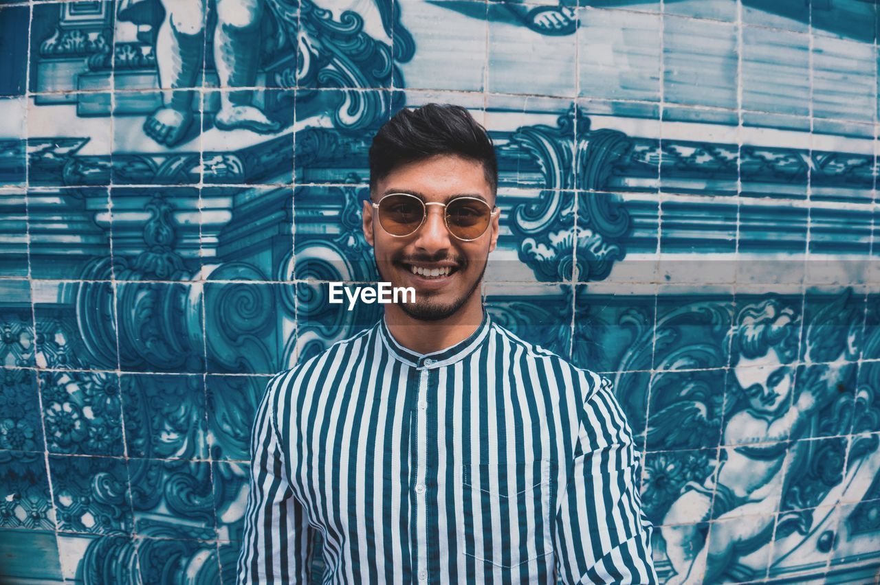 PORTRAIT OF YOUNG MAN WEARING SUNGLASSES STANDING AGAINST WALL