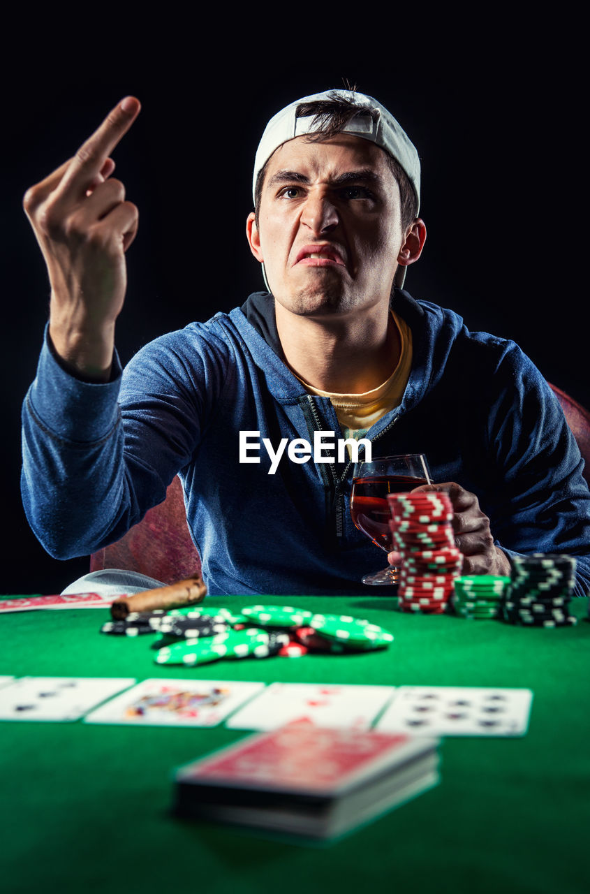 Young man showing obscene gesture while playing poker against black background