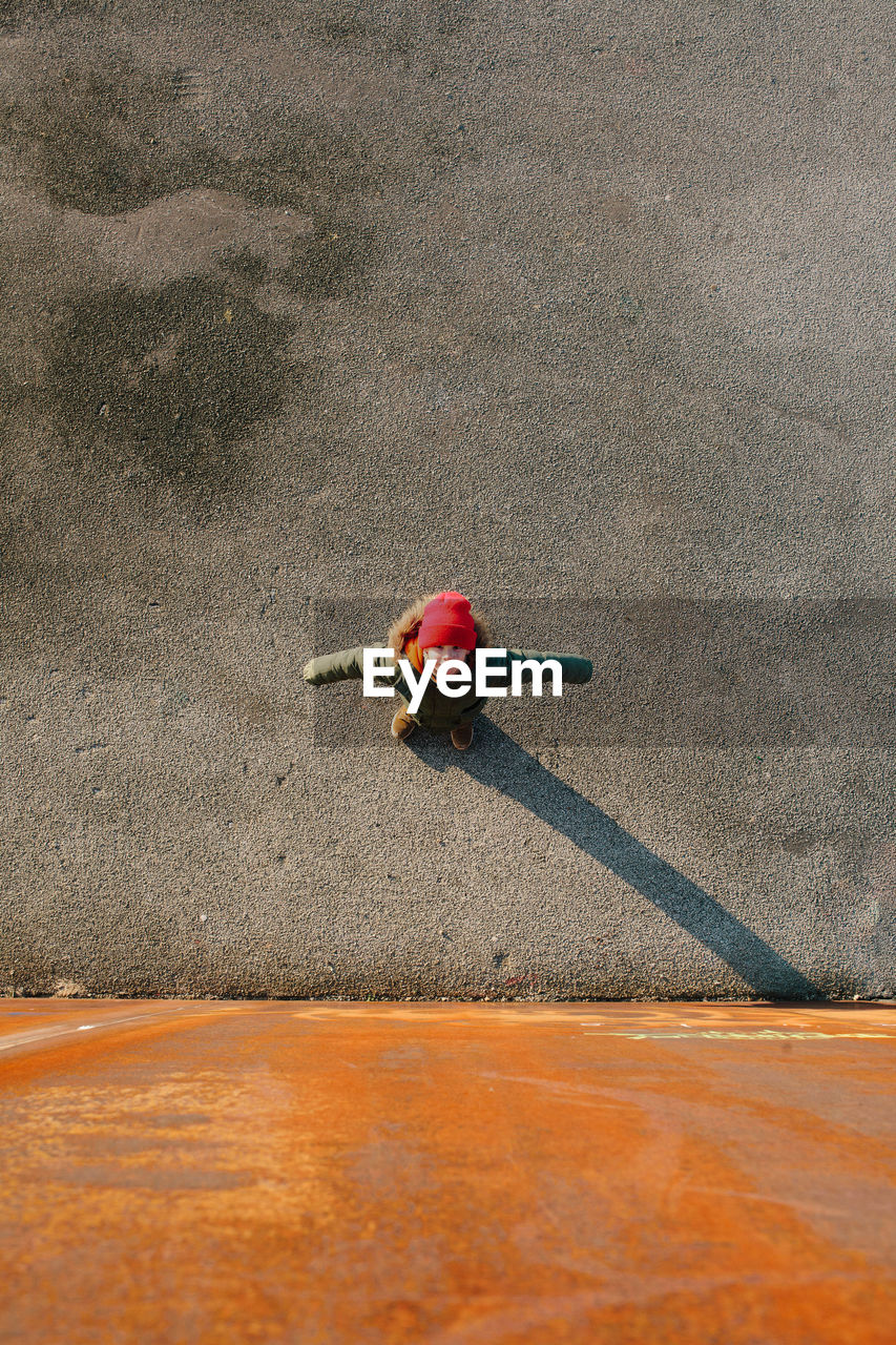 High angle view of girl standing with arms outstretched on street by orange wall