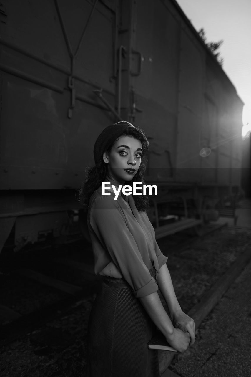 Dark haired female totally wearing clothes in vintage style standing near car train and holding open book in hands having dreaming look