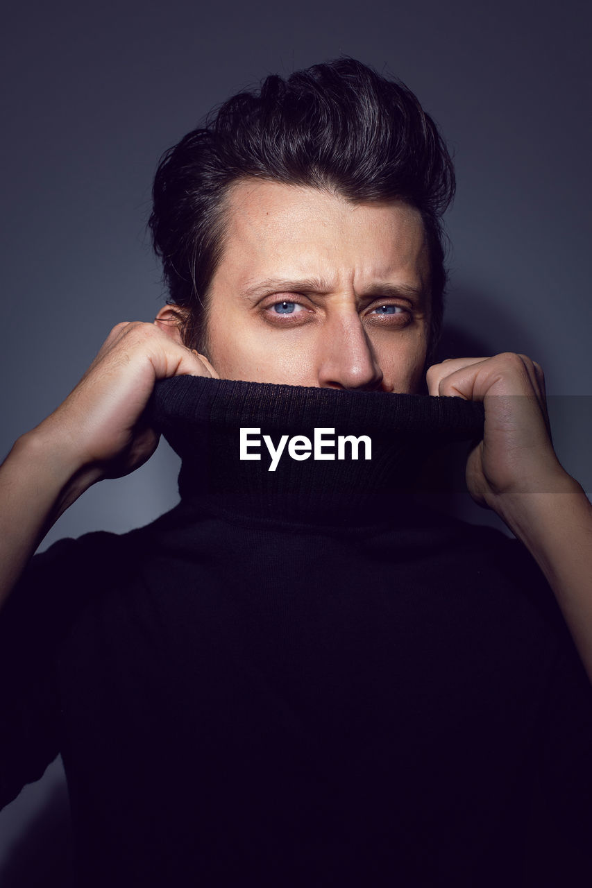 Portrait man standing over a light gray background in a black sweater covers his face. blue eyes