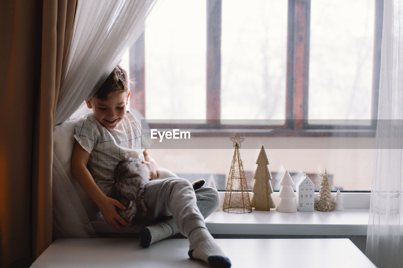 Cute boy playing with kitten sitting on the windowsill near the window. cozy home.