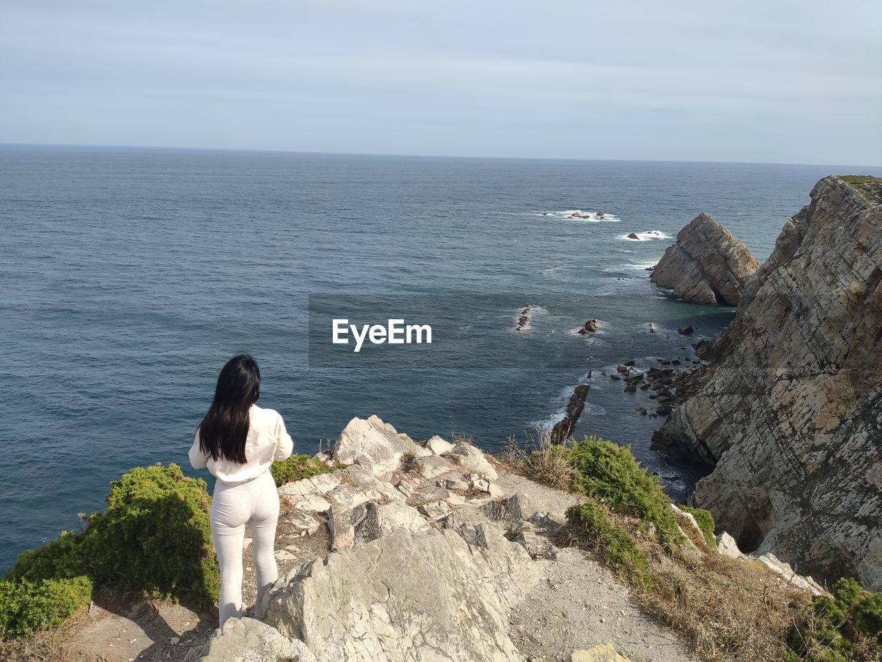 sea, water, rock, beauty in nature, horizon over water, horizon, cliff, scenics - nature, vacation, nature, sky, one person, leisure activity, shore, ocean, coast, tranquility, land, beach, lifestyles, women, day, rear view, tranquil scene, adult, terrain, non-urban scene, bay, full length, casual clothing, idyllic, rock formation, standing, outdoors, sitting, young adult, high angle view, sunlight, holiday, remote, looking at view, trip, relaxation