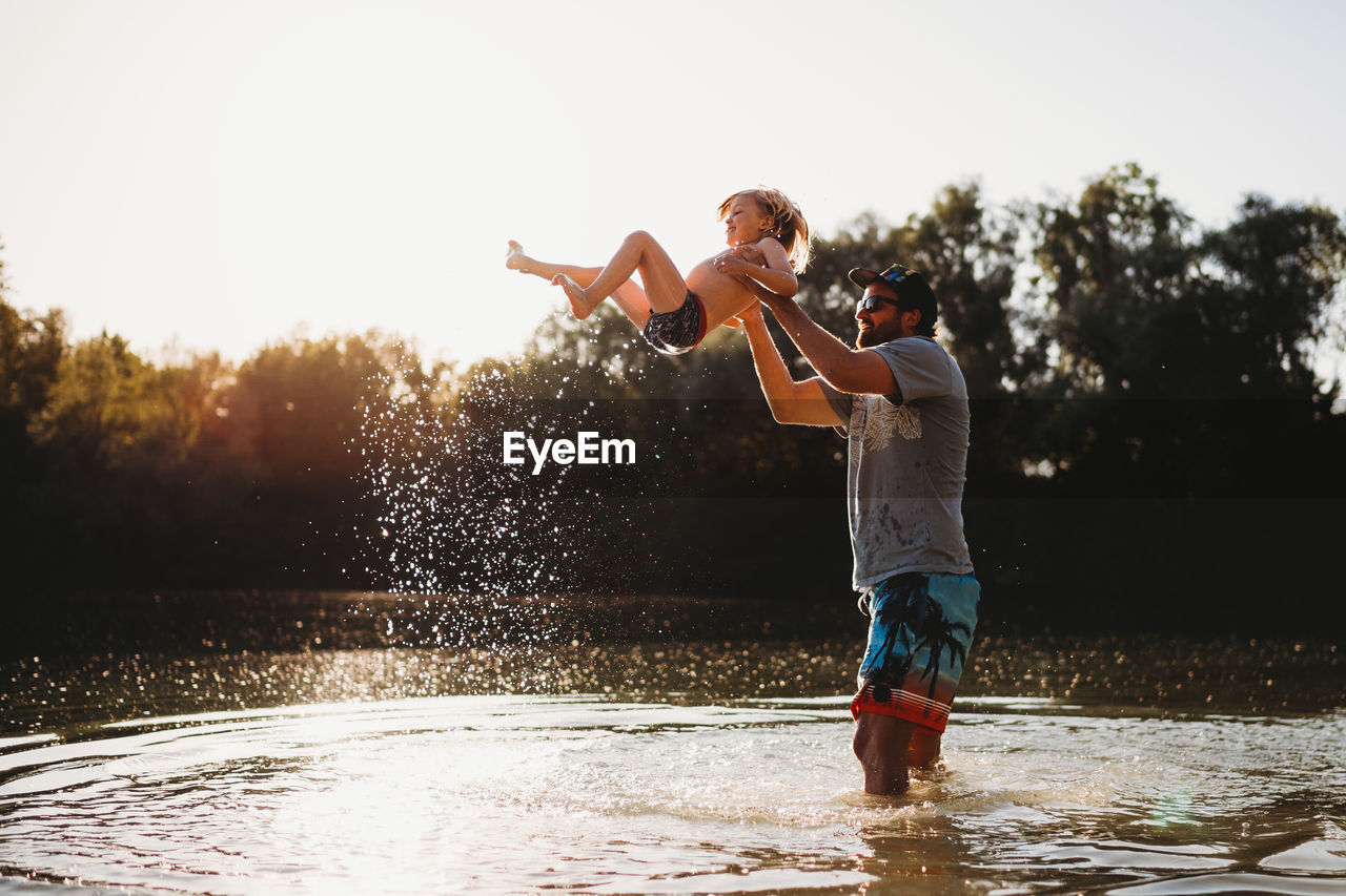 Father holding young child in the air at the lake splashing water