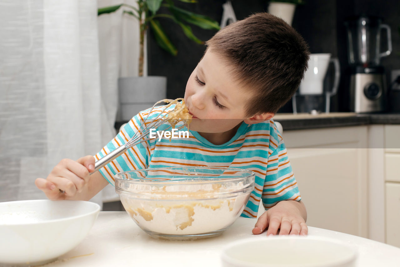 Cute boy licks a whisk with cake dough in the kitchen at home. time with children at home. 