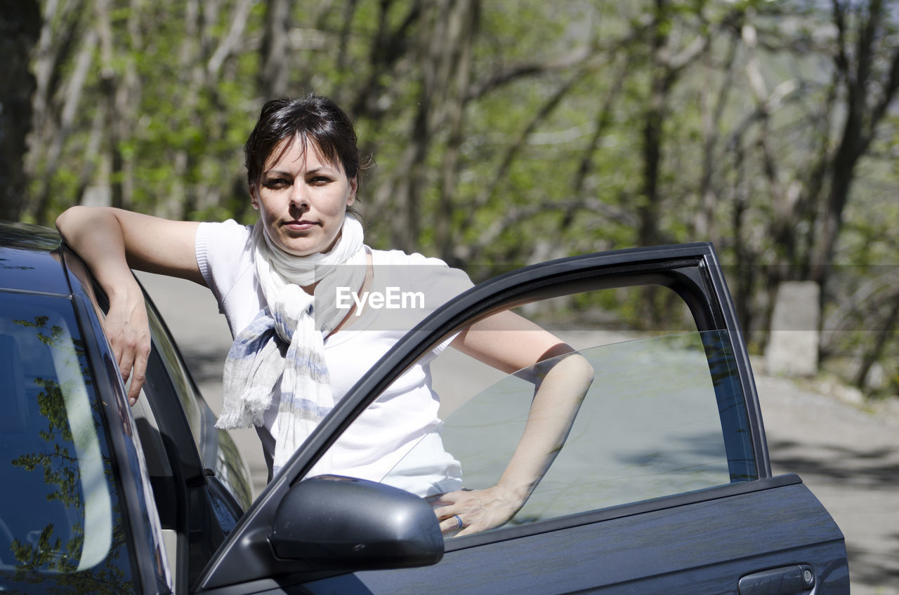 Portrait of confident woman standing by car against trees