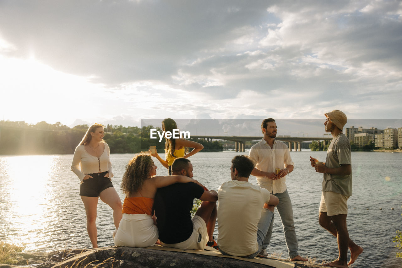 Multiracial male and female friends talking with each other during picnic near sea at sunset