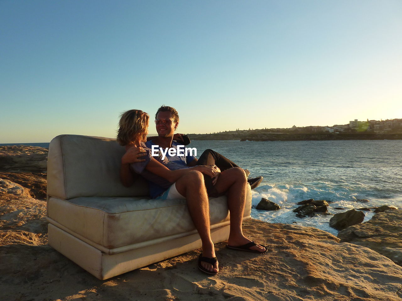 Couple sitting on sofa at beach against clear sky