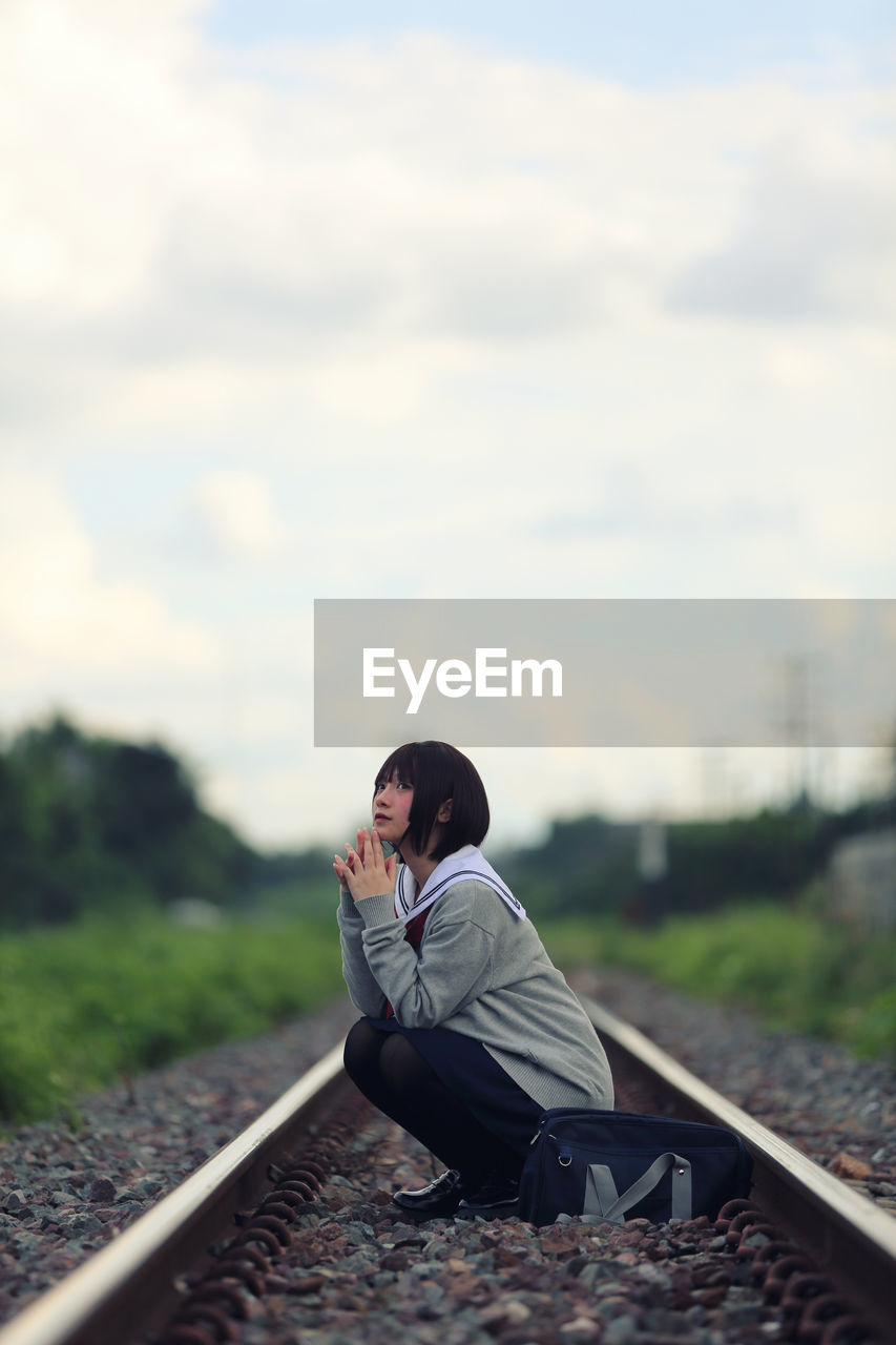 Young woman crouching on railroad track against sky