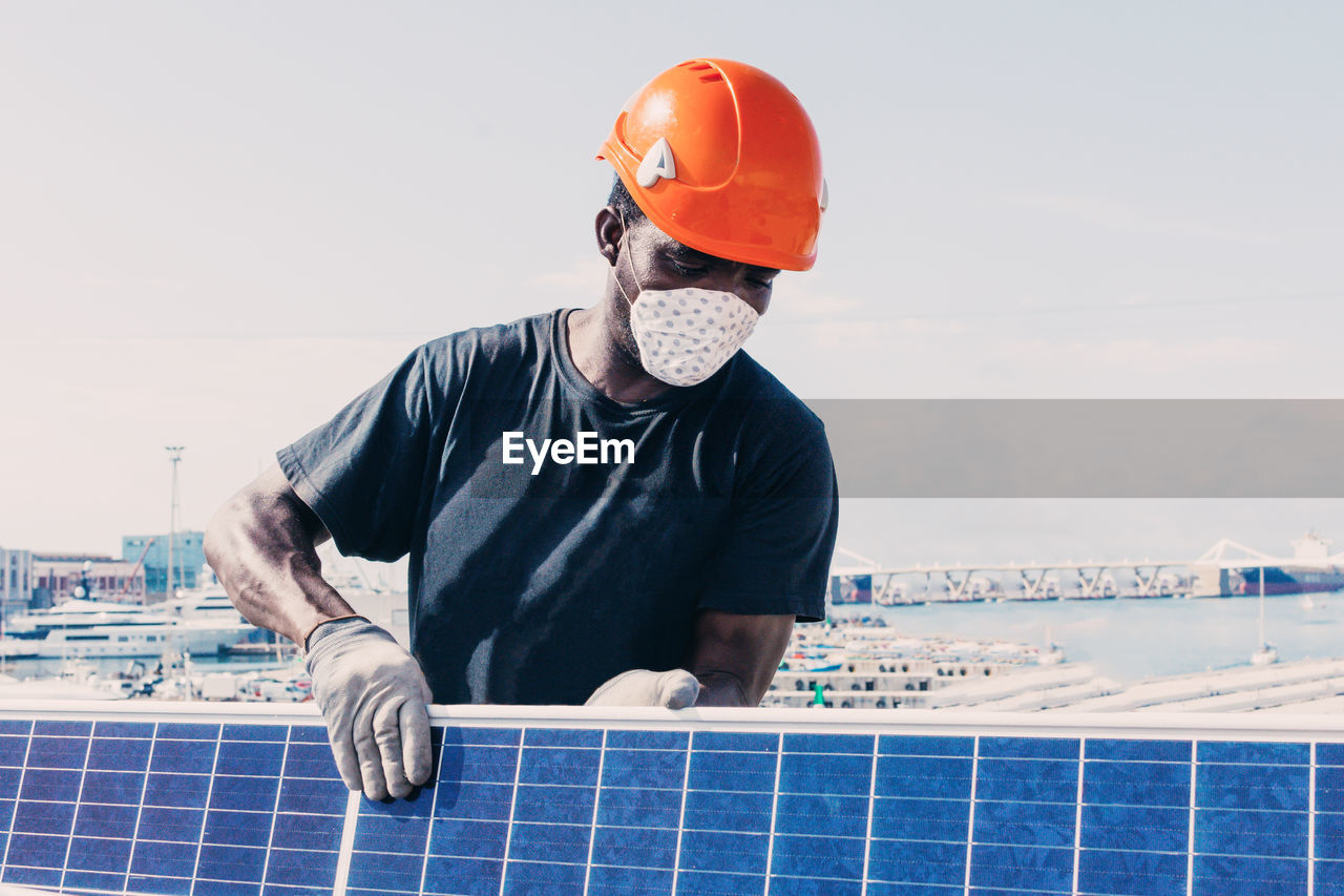 Black workman in helmet and protective mask installing solar battery in industrial area of contemporary plant