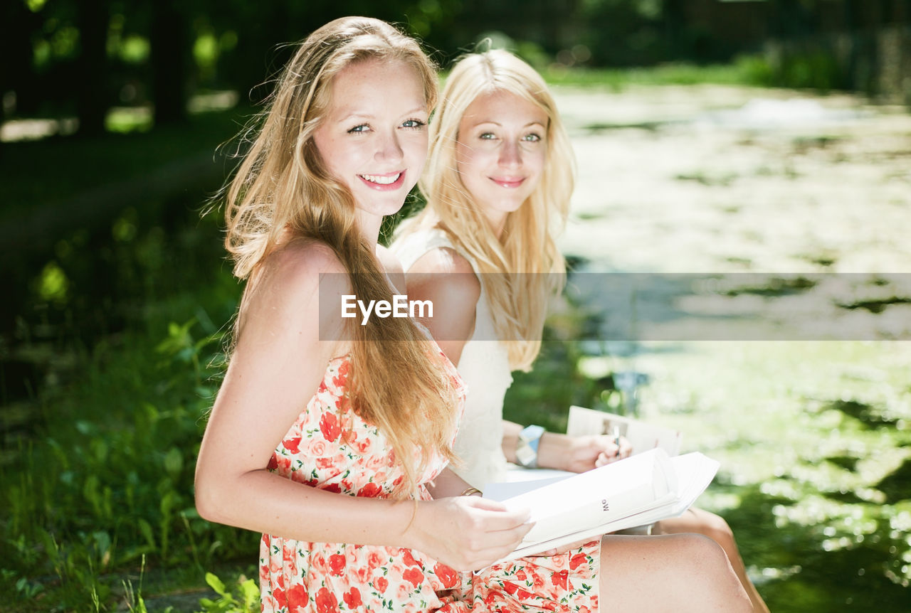 Portrait of smiling friends with books sitting in park during sunny day