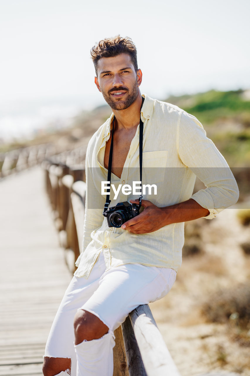 Portrait of man with camera sitting on railing against sky