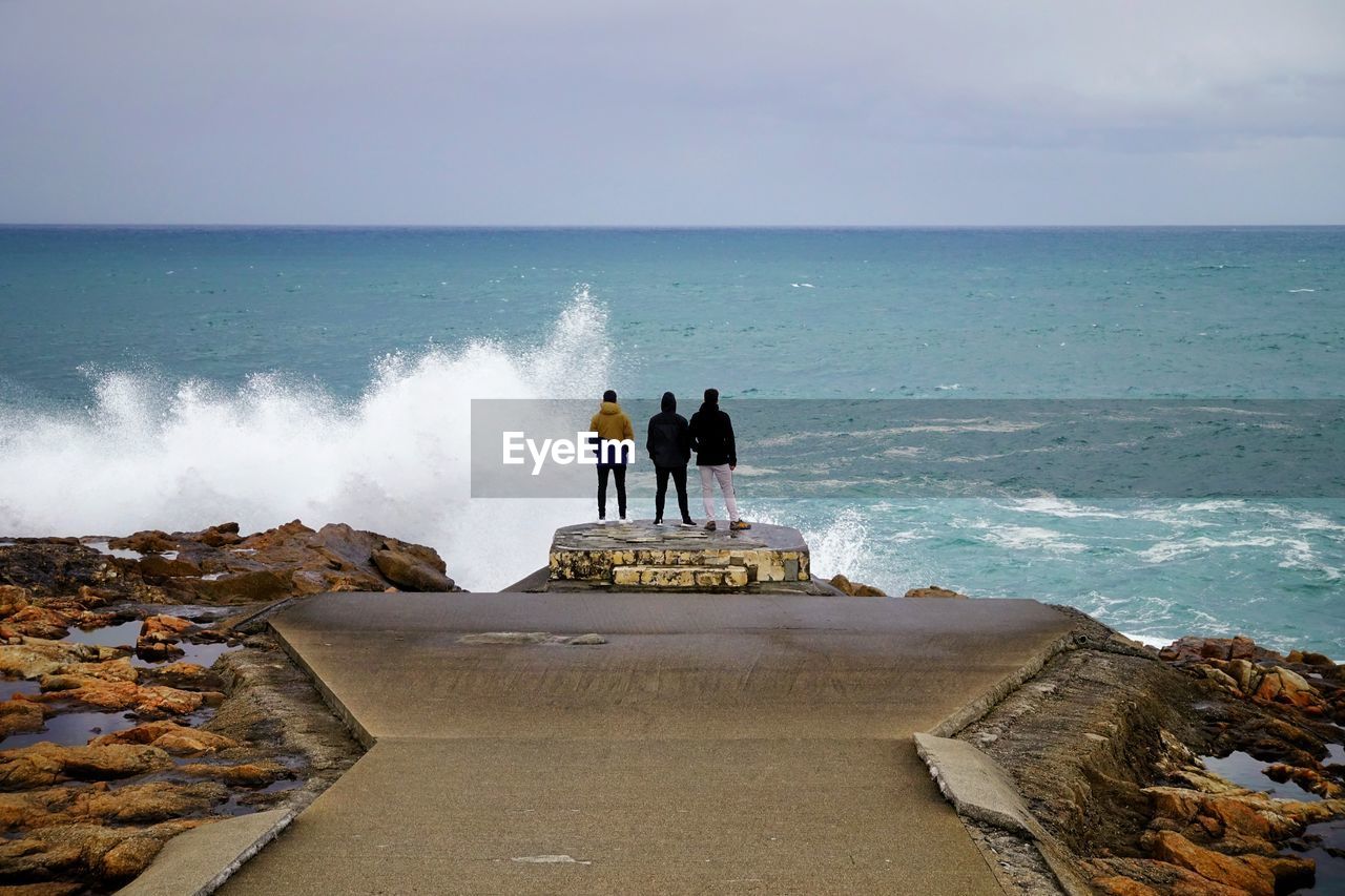 Rear view of men standing on cliff by sea