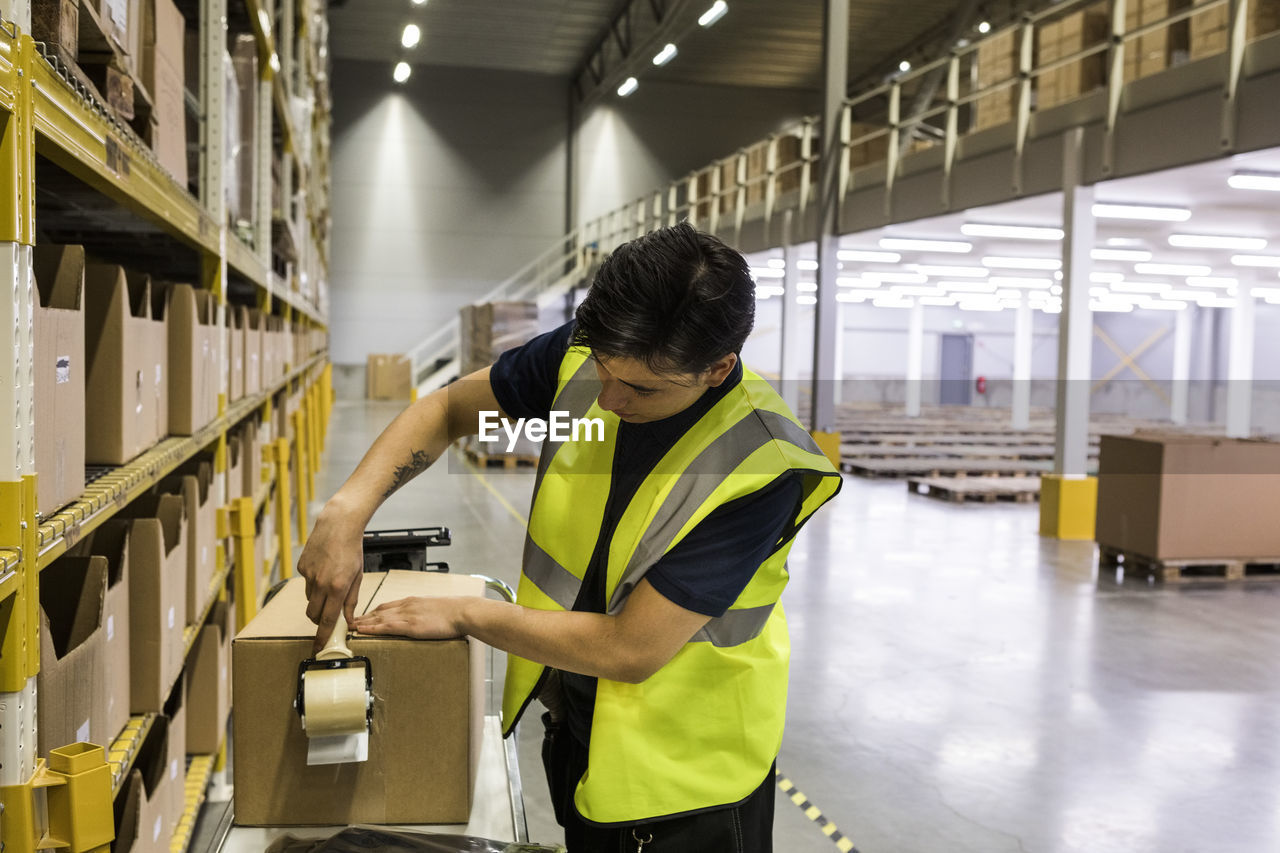 Young male worker packing cardboard box with adhesive tape at distribution warehouse