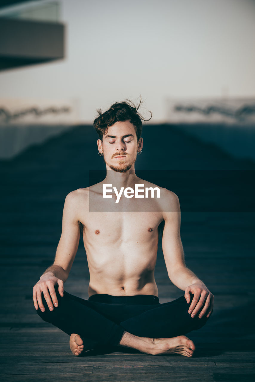 Shirtless young man meditating while sitting on boardwalk