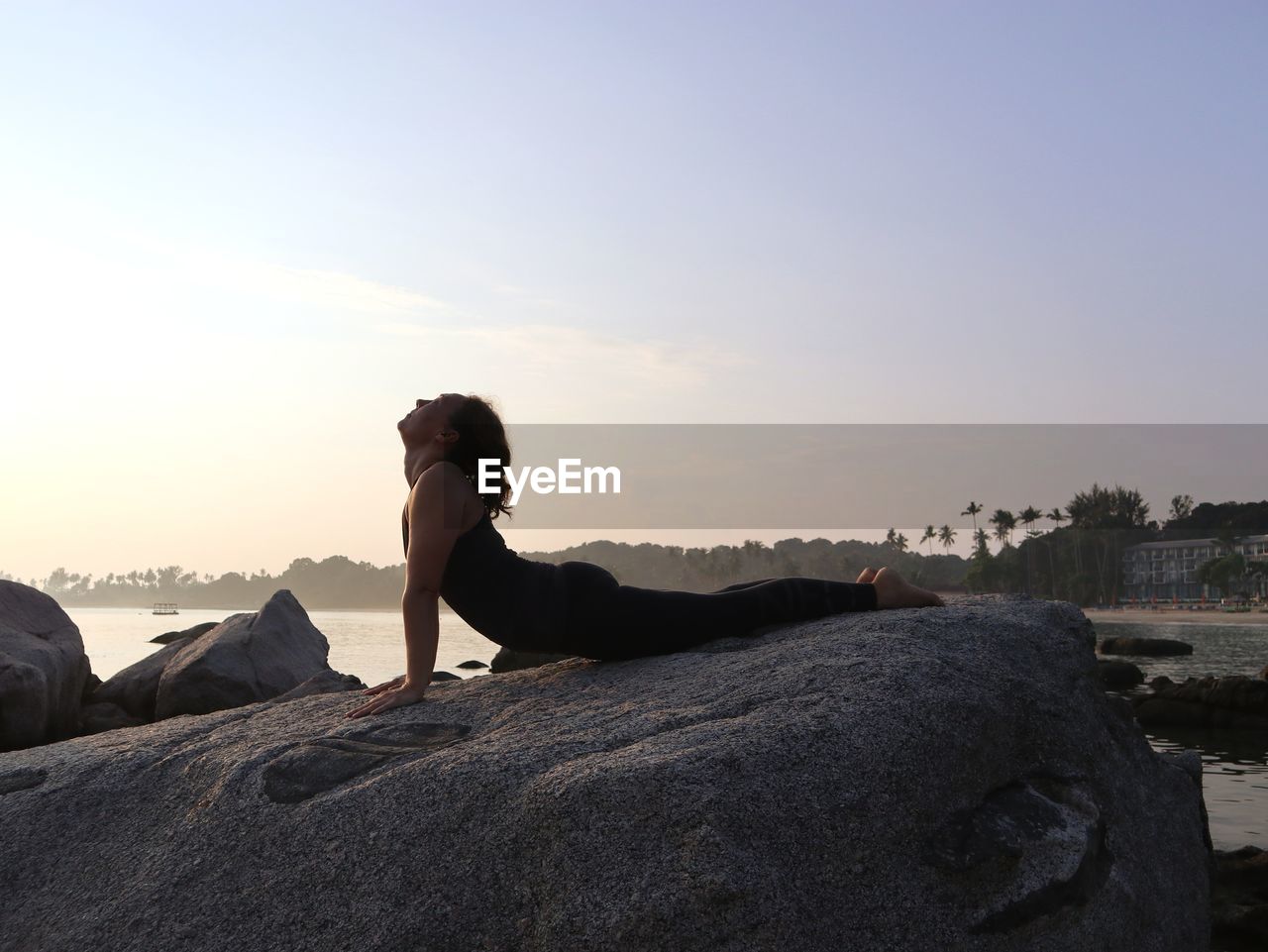 Woman practicing yoga, doing cobra pose on the rock, by the sea at sunrise