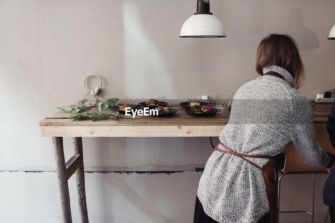 Rear view of female entrepreneur standing by table with food plates against wall at workshop