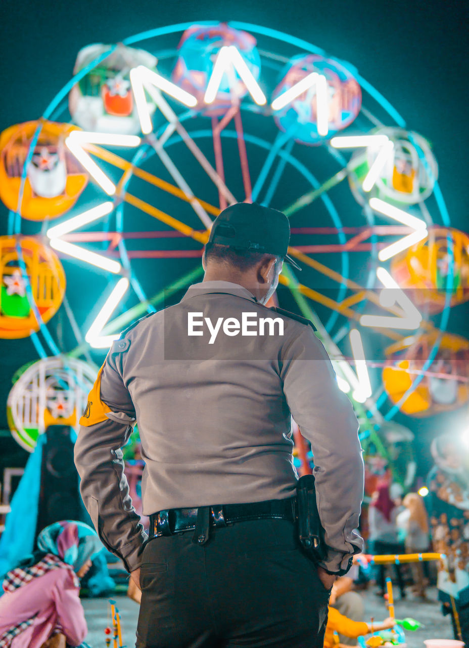 Rear view of man standing against illuminated ferris wheel at night
