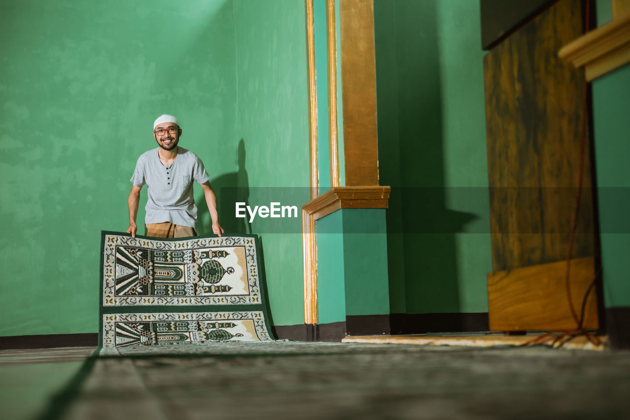 Happy young man with carpet standing against wall in mosque