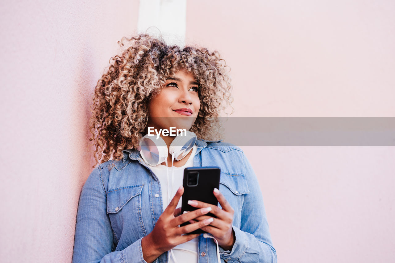 Portrait of smiling hispanic woman with afro hair in city using mobile phone and headset. lifestyle