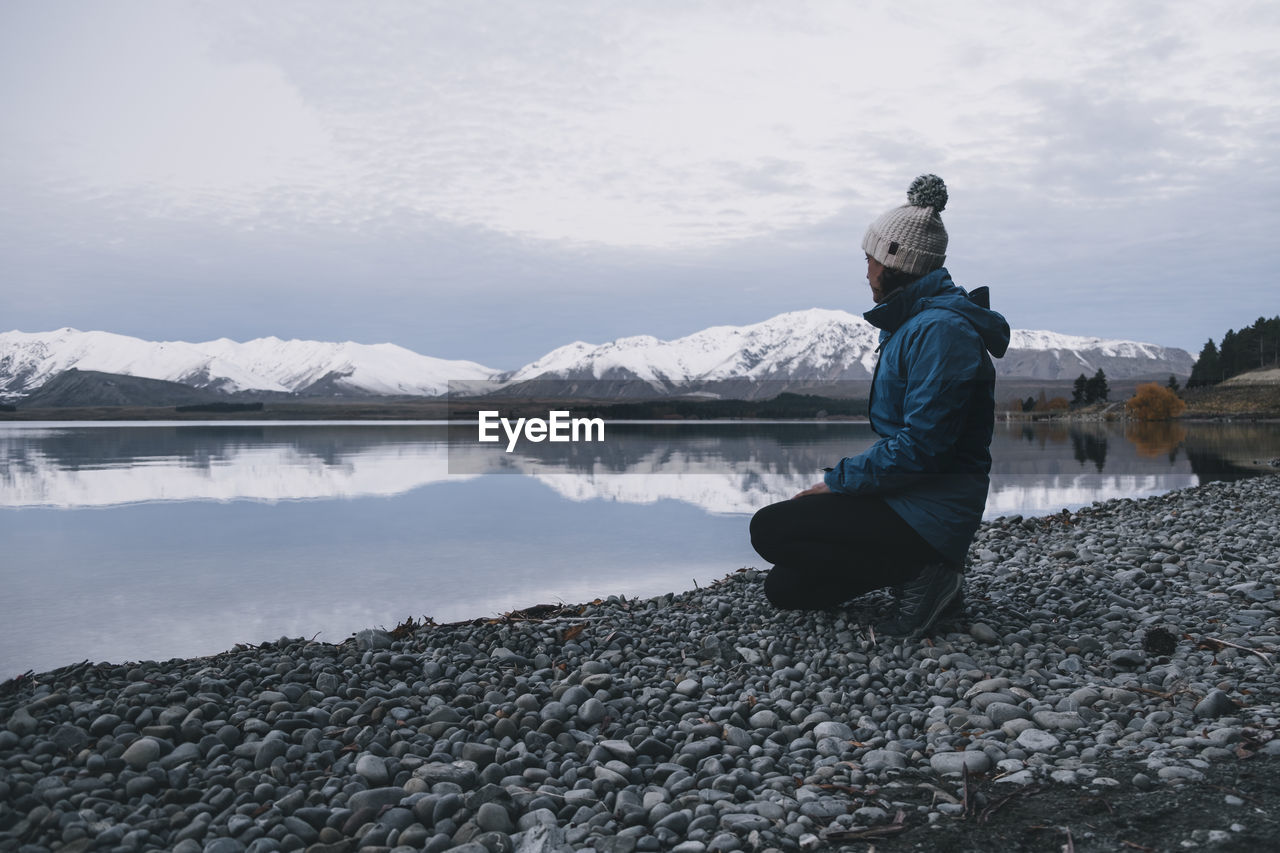 Young woman kneels down at lake tekapo and looks at the southern alps