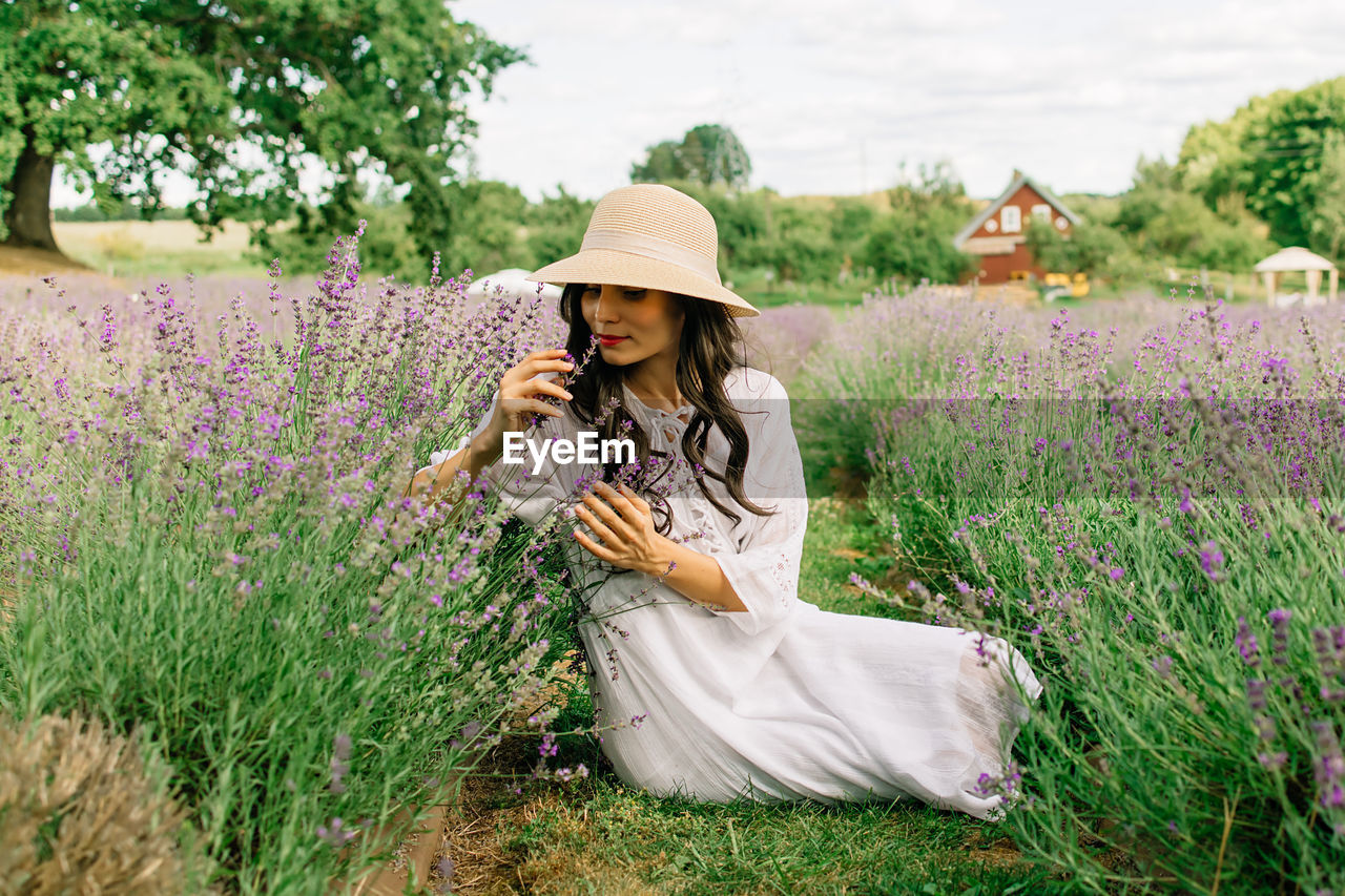 Close up of beautiful smiling woman in hat sitting on grass in lavender field 