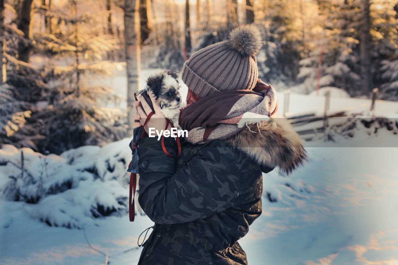 Side view of woman carrying puppy while standing on snow in forest during winter