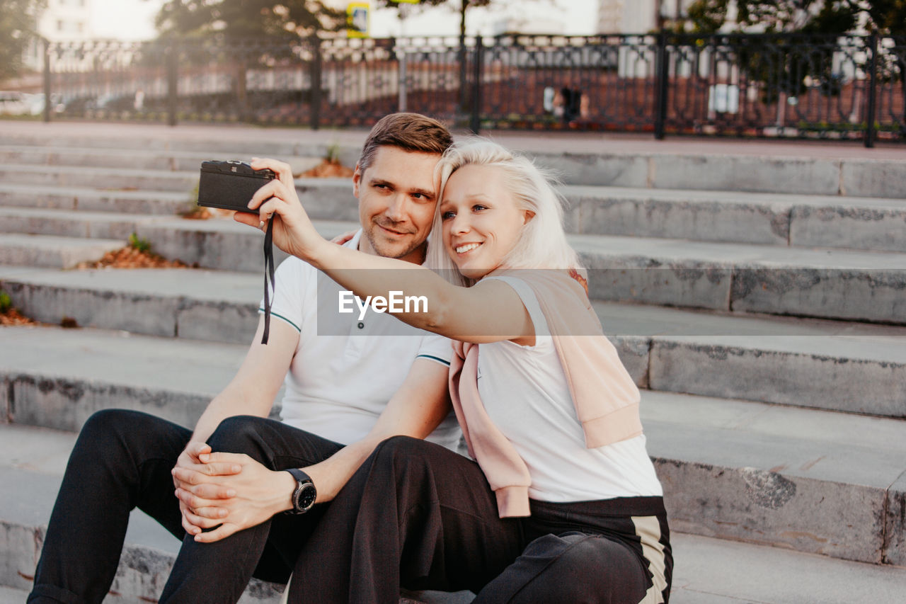Smiling couple taking selfie while sitting on staircase