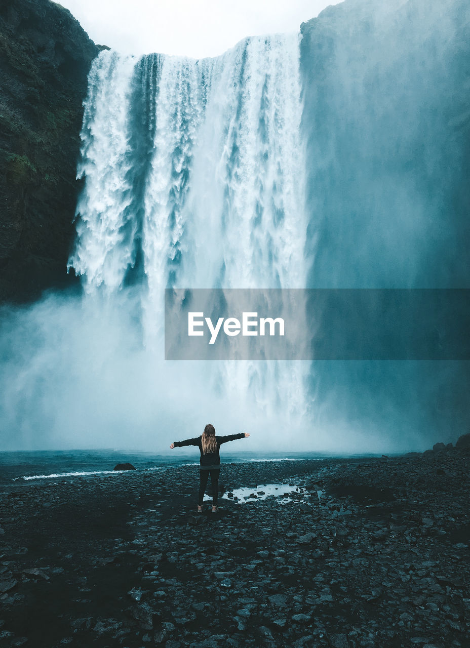 Rear view of woman with arms outstretched standing by waterfall