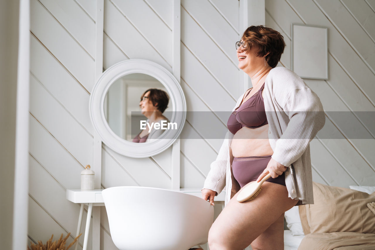 Middle age plus size woman in underwear doing body massage with dry wooden brush in bathroom at home