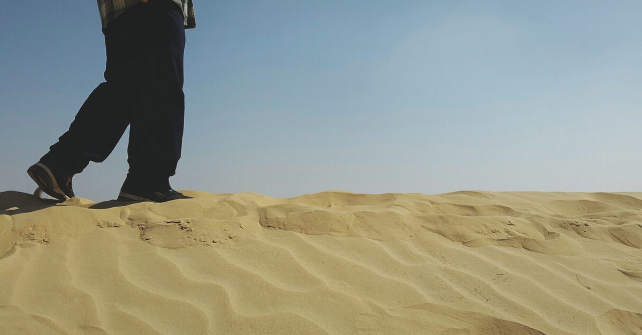 Low section of man walking on sand dune against clear sky